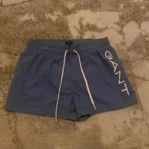 Blå badshorts från GANT - Snygga blå badshorts från GANT med vit snörning och stort tryck på sidan. Perfekta för en dag på stranden eller vid poolen, dem är aldrig använda och köptes för 1000kr från gants hemsida tveka inte på prisförslag 