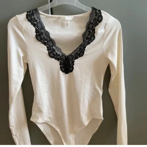 Vit långärmad body med spetsdetaljer - Säljer en vit body från H&M med långa ärmar och v-ringad spetsdetalj i svart. Perfekt vid alla tillfällen. Använd fåtal gånger!🥰❣️