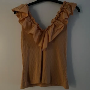 Orange topp med volang - Säljer en stilren orange ärmlös topp med volangdetaljer runt halsen. Toppen har en ribbad struktur som ger en fin textur 
