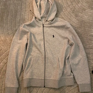 Grå hoodie från Ralph Lauren - Säljer en stilren grå hoodie från Ralph Lauren med dragkedja och broderad logga på bröstet. Tröjan har en klassisk design med huva och fickor framtill. Perfekt för en avslappnad look.