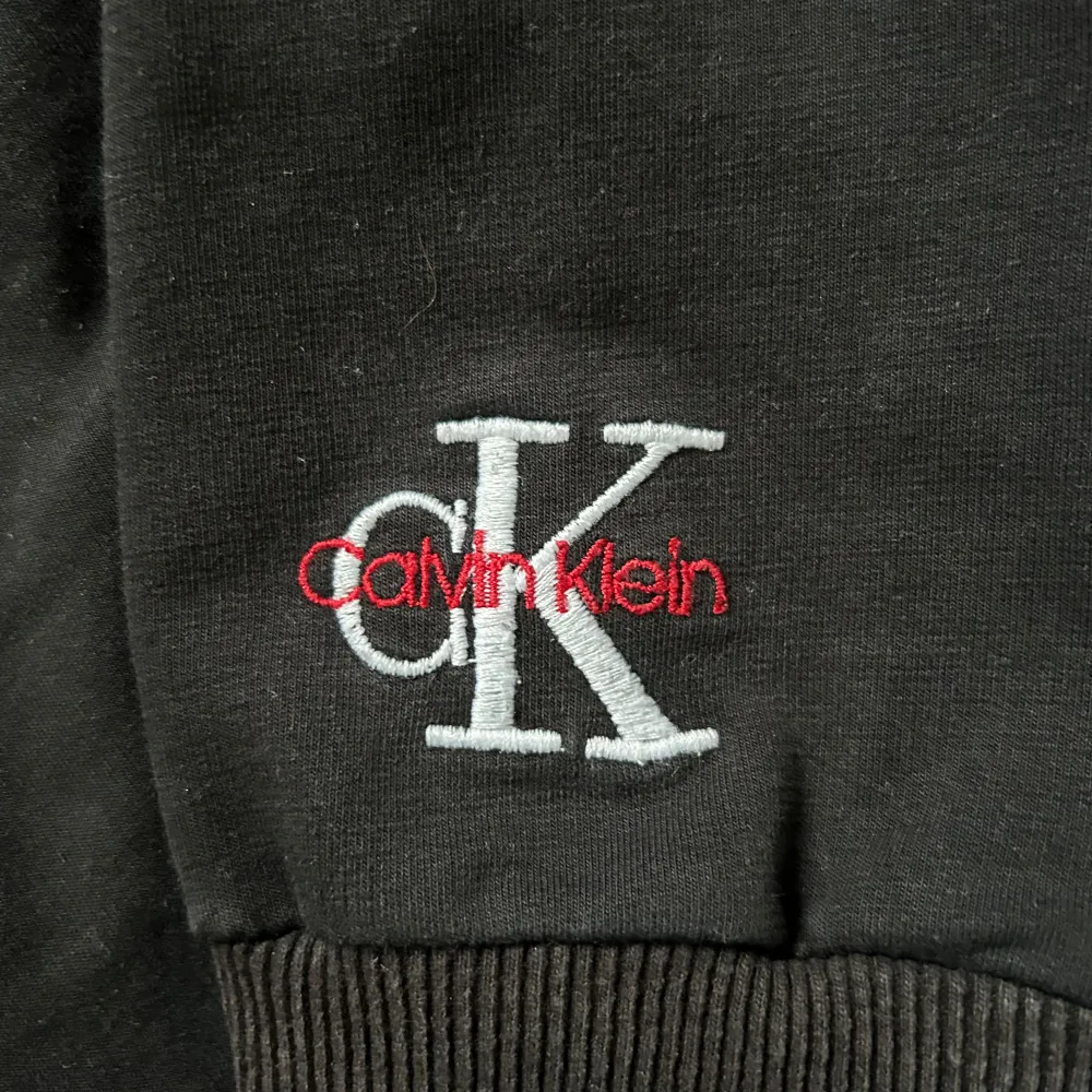 Snygg svart sweatshirt från Calvin Klein med det ikoniska CK-logotypen broderad i vitt och rött på bröstet. Tröjan har långa ärmar och ribbade muddar vid ärmslut och nederkant. Perfekt för en stilren och avslappnad look.. Neuletakit & Villapaidat.