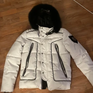 Vit pufferjacka från Horspist - Snygg vit pufferjacka från Horspist med svart foder och pälsdetaljer på huvan. Jackan har dragkedja och knappar framtill samt två praktiska fickor med dragkedja. Märkeslogga på ärmen ger en extra touch.