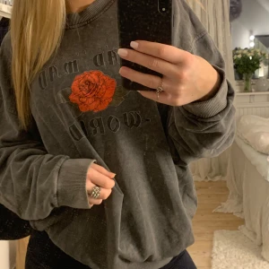Grå sweatshirt med rosdetalj - Säljer en grå sweatshirt med en stor röd ros och textdetaljer på både fram- och baksidan. Tröjan har en avslappnad passform och långa ärmar med ribbade muddar. Stlk S men passar större storlekar då den är oversized 