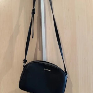 Svart axelväska från Calvin Klein  - Snygg svart axelväska från Calvin Klein med dragkedja upptill. Perfekt för att bära dina nödvändigheter med stil. Enkel och elegant design som passar till många tillfällen.