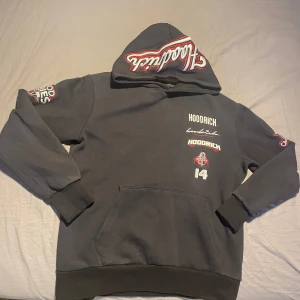 Svart hoodie från Hoodrich - Snygg svart hoodie från Hoodrich med tryck i vitt och rött. Den har en stor ficka framtill och textdetaljer på ärmarna och ryggen. Perfekt för en avslappnad stil.