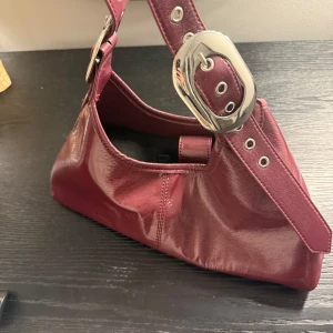 Vinröd axelväska med stora spännen - Snygg vinröd axelväska i glansigt material med stora metallspännen. Kommer faktiskt inte ihåg vart jag köpte, men märket är topshop och har använts max 3 ggr 