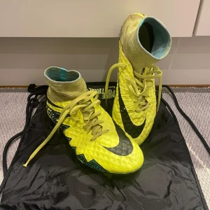 Nike Hypervenom Elite SG - Snygga gula fotbollsskor från Nike med hög ankelkrage och snörning. Skorna har en iögonfallande design med svarta och turkosa detaljer samt en robust sula med dobbar för bra grepp på planen. Värt att veta: innersulan i vänster sko är så pass sliten att den lossnat lite. Skickar bild vid behov.