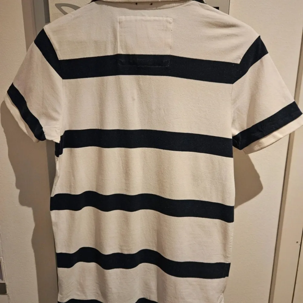 Säljer en snygg vit och svart randig pikétröja från Abercrombie & Fitch. Tröjan har korta ärmar och en klassisk krage med knappar. Den har en cool broderad detalj på bröstet.. T-paidat.