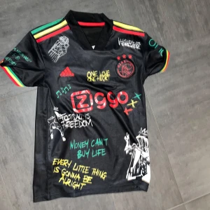Svart fotbollströja med Bob Marley-tema - Cool svart fotbollströja från Adidas med Bob Marley-tema. Tröjan har färgglada detaljer i rött, gult och grönt på ärmarna och kragen. Den är dekorerad med olika tryck och citat, inklusive en stor bild av Bob Marley på baksidan. Perfekt för fotbollsfans som älskar reggae!