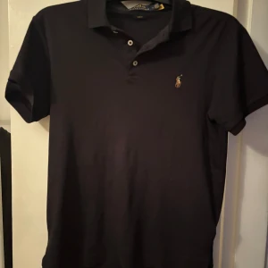 Svart pikétröja från Polo Ralph Lauren - Snygg svart pikétröja från Polo Ralph Lauren i slim fit. Tröjan har korta ärmar och en klassisk krage med knappar. Perfekt för en stilren look.