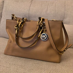 Beige handväska från Michael Kors - Snygg beige handväska från Michael Kors med gulddetaljer och logotyp. Väskan har både handtag och en axelrem. Insidan är rymlig med flera fack och dragkedja. Har använts endast några få gånger💗pris kan diskuteras!