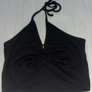 Svart halterneck topp - Snygg svart halterneck topp med knytning i nacken och en stilren design. Toppen har en omlott-look framtill med en dekorativ rynkning i mitten. Perfekt för en trendig och avslappnad stil.
