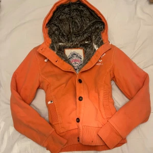 Orange jacka med pälsfoder från Geographical Norway - Säljer en snygg orange jacka från Geographical Norway med mjukt fuskpälsfoder i luvan. Jackan har knappar framtill och ribbade muddar vid ärmslut och nederkant. Perfekt för kyliga dagar! Vintage därav priset. Storlek m men sitter som s/xs