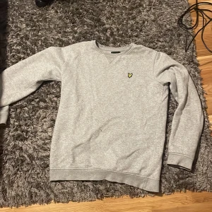 Grå sweatshirt från Lyle & Scott - Säljer en stilren grå sweatshirt från Lyle & Scott med deras ikoniska gula logga på bröstet. Tröjan har långa ärmar och en rund halsringning. Perfekt för en avslappnad look.