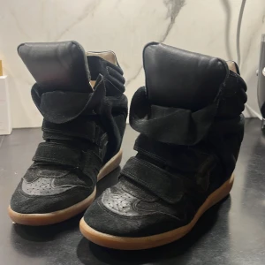 Svarta skor från Isabel Marant - Snygga svarta sneakers från Isabel Marant med hög design och kardborreband. Skorna har en robust gummisula och är tillverkade i mocka och skinn. Perfekta för en trendig look. Knapppt använda 