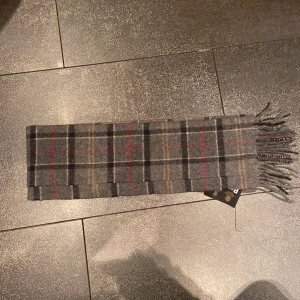 Grå rutig halsduk från Barbour - Snygg grå rutig halsduk från Barbour i 100% ull. Den har ett klassiskt rutmönster med inslag av rött och beige, perfekt för att hålla dig varm under kyliga dagar. Fransen på kanterna ger en stilren touch.