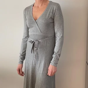 Esprit Klänning - Elegant grå klänning från Esprit med v-ringning och långa ärmar. Klänningen har ett knytband i midjan och en plisserad kjol som ger en fin siluett. Perfekt för både vardag och speciella tillfällen. Modellen är 176 cm lång.