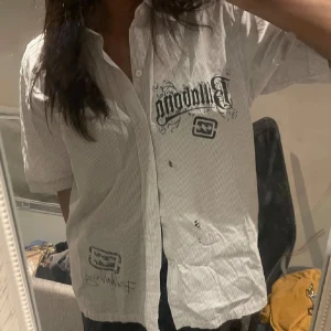 Vit kortärmad skjorta med tryck - Säljer en vit kortärmad skjorta med tryck framtill. Skjortan har en avslappnad passform och knappar längs framsidan. Perfekt för en casual look.