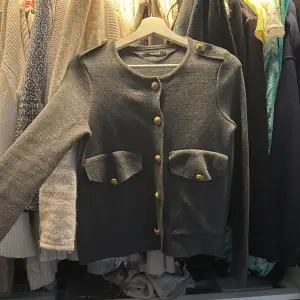 Snygg grå kofta från Gina Tricot med guldknappar och dekorativa fickor framtill. Koftan har en rund halsringning och långa ärmar, perfekt för en stilren look.