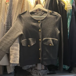 Grå kofta  - Snygg grå kofta från Gina Tricot med guldknappar och dekorativa fickor framtill. Koftan har en rund halsringning och långa ärmar, perfekt för en stilren look.