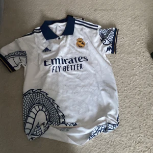 Vit Real Madrid fotbollströja från Adidas - Säljer en vit Real Madrid fotbollströja från Adidas med blå detaljer och klubbens emblem på bröstet. Tröjan har korta ärmar och ett unikt drakmönster. Perfekt för alla fans av laget!