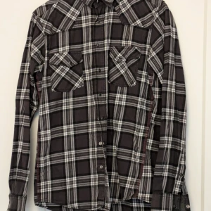 Grå rutig skjorta - Snygg svart och vit rutig skjorta med långa ärmar och knappar framtill. Skjortan har två bröstfickor med lock och är tillverkad i bomull med en liten del elastan för extra komfort. Perfekt för en avslappnad stil. 