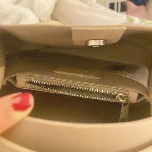 Beige handväska med dragkedja - Elegant beige handväska med rundad form och praktisk dragkedja inuti. Väskan har en enkel design med en knappstängning och ett bekvämt handtag för enkel bärning. Perfekt för att hålla dina saker organiserade och stilrena.