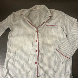 Randig pyjamasskjorta från Body Zone - Mysig randig pyjamasskjorta från Body Zone i ljusblått och vitt med röda knappar och detaljer. Skjortan har en bröstficka och långa ärmar, perfekt för en avslappnad kväll. Den är använd 