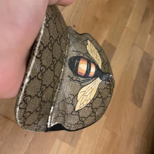 Gucci bi - Snygg keps från Gucci med ett ikoniskt mönster och en broderad bi på framsidan. Kepsen har justerbart band med gröna och röda ränder. Perfekt för att ge din outfit en lyxig touch.