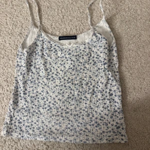 Blommig linnetopp från Brandy Melville - Söt blommig linnetopp från Brandy Melville i vitt och blått. Toppen har smala axelband och en liten rosett framtill. Perfekt för varma sommardagar!