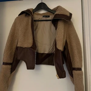 Beige teddyjacka  - Säljer en snygg beige teddyjacka från  med bruna detaljer. Jackan har en croppad passform och dragkedja framtill. Perfekt för kyliga dagar när du vill vara både varm och trendig.