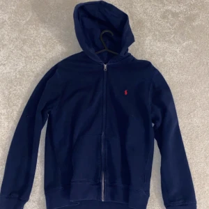 Mörkblå hoodie från Ralph Lauren - Snygg mörkblå hoodie från Ralph Lauren med dragkedja och huva. Den har en liten röd logga på bröstet och är perfekt för en avslappnad stil. Passar bra till både jeans och träningsbyxor.