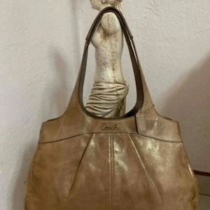 Beige/ Golden  axelväska från Coach - Snygg beige axelväska från Coach  med glansig finish. Väskan har två långa handtag och en praktisk dragkedja upptill. Insidan är rymlig med flera fack för enkel organisering. 