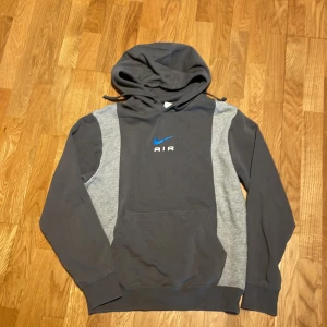 Grå och blå hoodie från Nike - Säljer en snygg grå hoodie från Nike med blått 'Air'-tryck på bröstet. Tröjan har en klassisk känguruficka och justerbar huva med dragsko. Perfekt för en avslappnad stil.