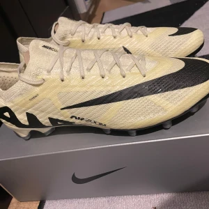 Nike Air Zoom fotbollsskor - Snygga Nike Air Zoom fotbollsskor i beige och svart. Skorna har en modern design med snörning och är perfekta för fotbollsplanen. De är tillverkade i syntetmaterial för optimal prestanda.