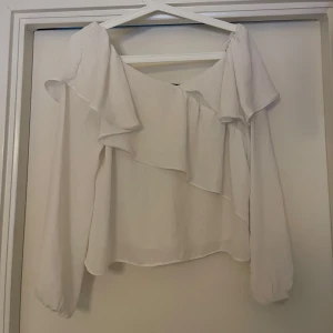 Vit blus från Gina Tricot - Säljer en elegant vit blus från Gina Tricot med volangdetaljer. Blusen har långa ärmar och en luftig passform, perfekt för en stilren look.