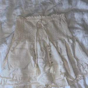 Beige kjol från Zara - Charmig beige kjol från Zara med smockdetaljer och volangkant. Kjolen har en knytning framtill med tofsar och är perfekt för en somrig look. Även super fin att använda som topp!😇