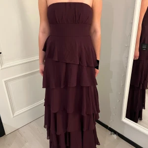 Vinröd axelbandslös klänning - Elegant vinröd axelbandslös klänning med flera lager volanger som ger en vacker rörelse. Perfekt för speciella tillfällen. Klänningen har en dragkedja i ryggen för enkel påklädning. Aldrig använd och prislappen är kvar, storlek S!💗