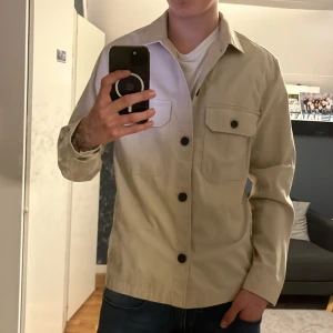 Beige overshirt från H&M - Säljer en stilren beige overshirt från H&M med knappar framtill och två bröstfickor. Perfekt för lager-på-lager-stil och passar till många olika outfits.