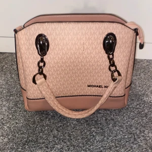 Rosa handväska från Michael Kors - Snygg rosa handväska från Michael Kors med logotypmönster. Väskan har svarta detaljer och robusta handtag. Perfekt för att bära dina dagliga nödvändigheter med stil.