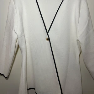 Vit omlottkavaj från Zara - Elegant vit omlottkavaj från Zara med svarta kanter och en knappdetalj framtill. Kavajen har 3/4 långa ärmar och en stilren design som passar perfekt för en chic look.