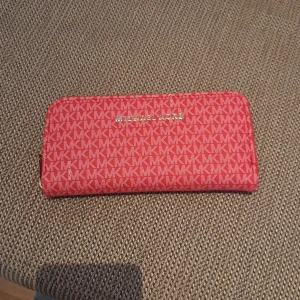 Röd plånbok från Michael Kors (falsk)  - Snygg röd plånbok från Michael Kors (falsk) med det klassiska MK-mönstret. Plånboken har en dragkedja och flera fack för kort och kontanter. Perfekt för att hålla ordning på dina tillhörigheter med stil.