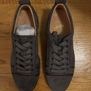 Grå sneakers från Christian Louboutin - Snygga grå sneakers i mocka från Christian Louboutin med den ikoniska röda sulan. Skorna har snörning och en stilren design som passar perfekt för en trendig look. Aldrig använda och är storlek 41