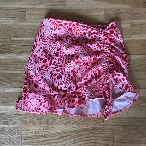 Rosa och röd mönstrad kjol med volang - Säljer en snygg kortkjol i rosa och rött med ett djärvt leopardmönster. Kjolen har en volangdetalj längst ner och snörning på sidan för en justerbar passform. Perfekt för att sticka ut med stil!