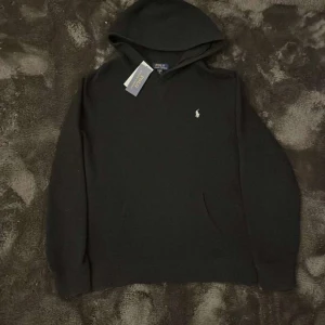 Svart hoodie från Ralph Lauren - Säljer en stilren svart hoodie från Ralph Lauren med en liten broderad logga på bröstet. Perfekt för en avslappnad look. Hoodien har långa ärmar och en klassisk huva.