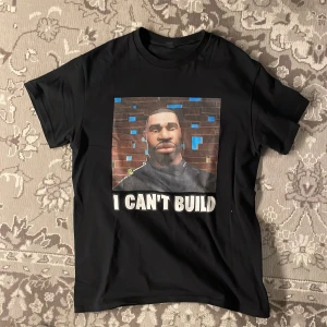 Svart t-shirt med tryck - Snygg svart t-shirt med ett humoristiskt tryck av en karaktär framför en tegelvägg och texten 'I CAN'T BUILD'. Perfekt för gaming-entusiaster eller som en rolig vardagströja.