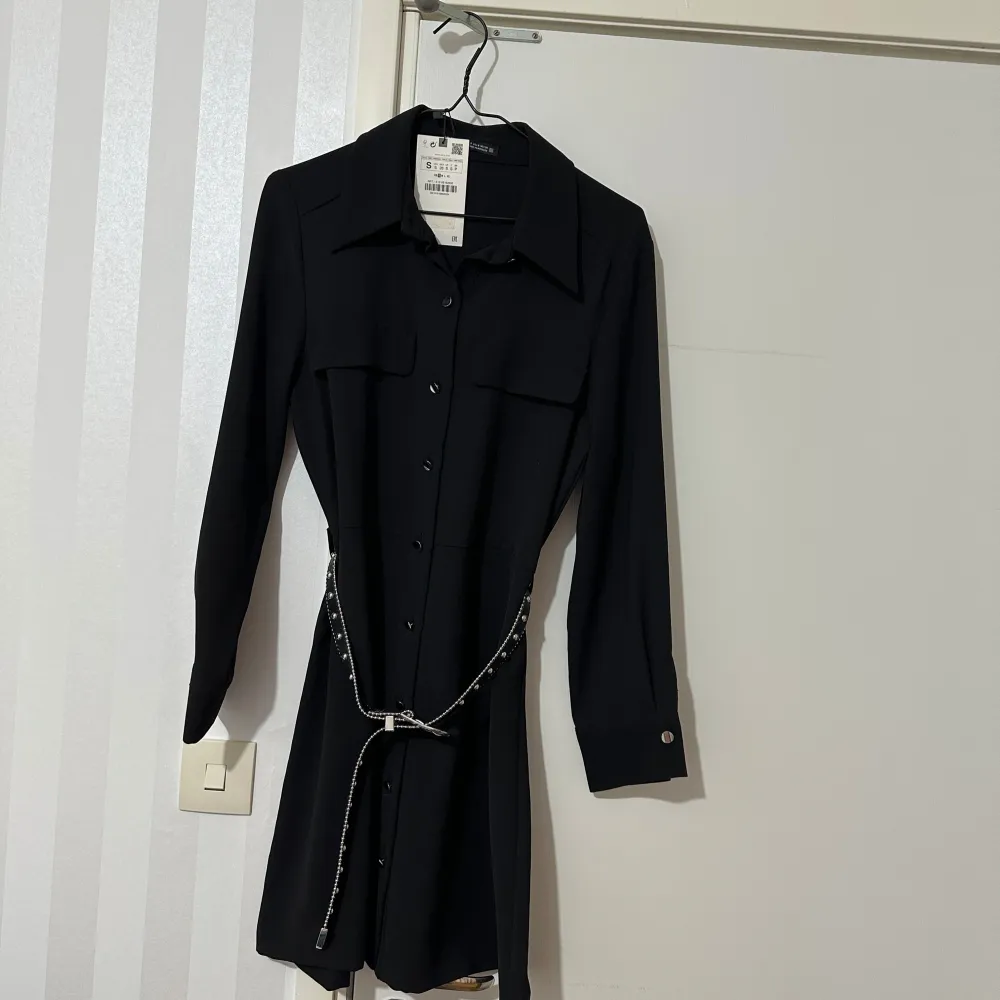 Elegant svart klänning med långa ärmar och knappar framtill. Den har en stilren krage och två bröstfickor. Klänningen kommer med ett glittrigt bälte som ger en extra touch av glamour.. Mekot.