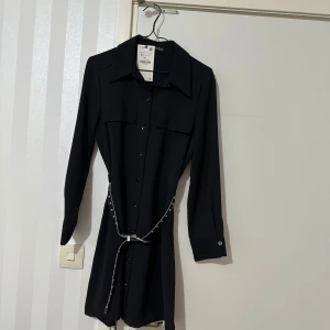 Svart klänning med bälte - Elegant svart klänning med långa ärmar och knappar framtill. Den har en stilren krage och två bröstfickor. Klänningen kommer med ett glittrigt bälte som ger en extra touch av glamour.