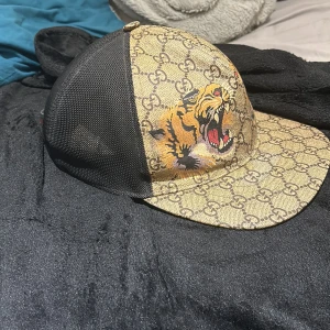 Mönstrad keps med tigertryck - Snygg keps med ett djärvt tigertryck på framsidan och ett klassiskt mönster. Kepsen har en svart mesh-baksida och justerbart band i rött och grönt. Perfekt för att ge din outfit en unik touch väldigt sällsynt och äkta som ni ser stolek xl men har m själv och passar perfekt 