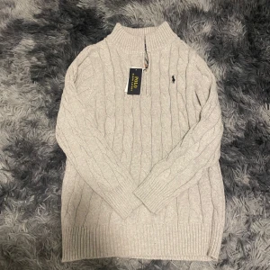 Grå stickad tröja från Polo Ralph Lauren - Säljer en grå stickad tröja från Polo Ralph Lauren med kabelstickat mönster. Tröjan har långa ärmar och en hög krage. Dessutom oanvänd. Hör av er vid frågor! EJ ORIGINAL 